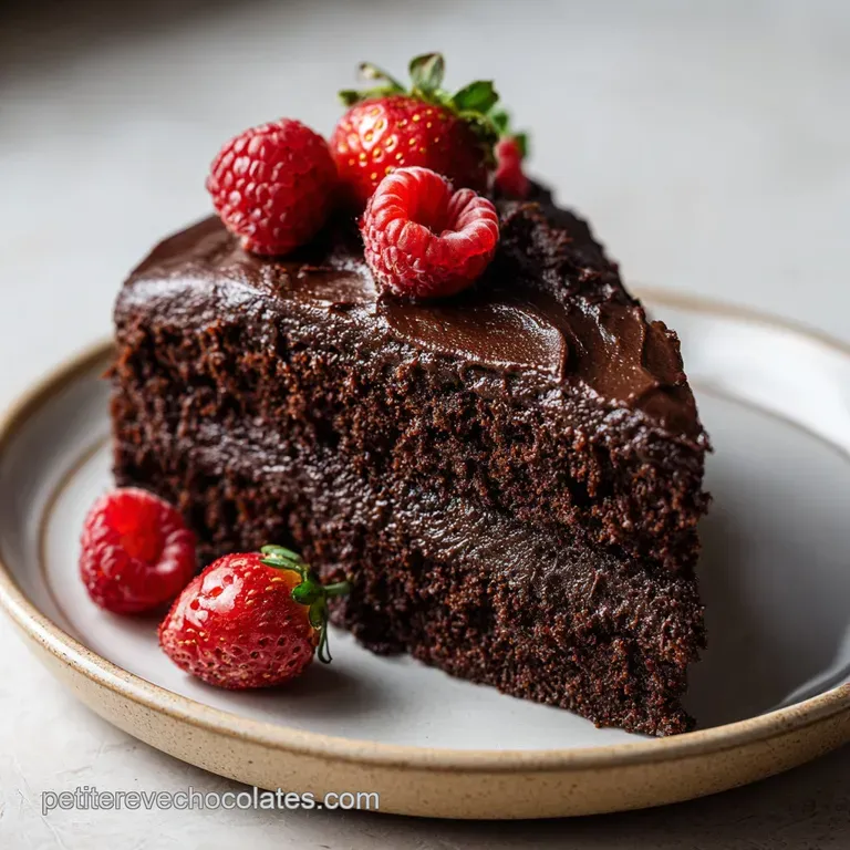 A slice of moist chocolate cake on a white plate, topped with a dollop of whipped cream and a raspberry.